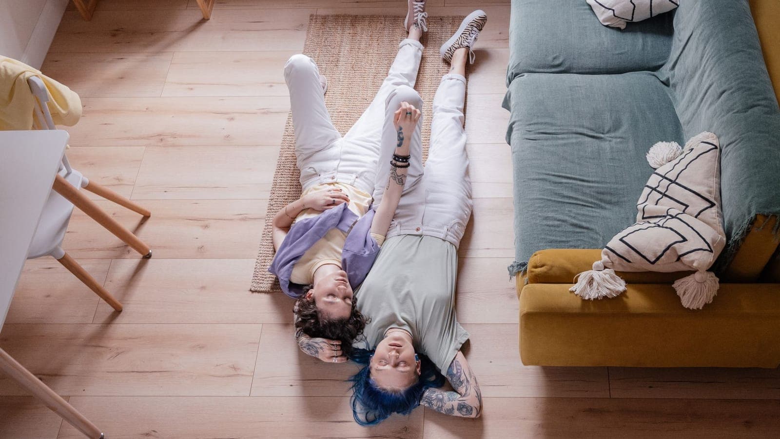 Two people dressed in white lying on the wooden floor of a living room, seen from above, next to a yellow sofa covered with a green throw
