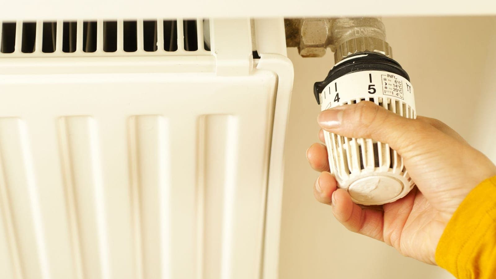 A hand in a yellow sleeve turning the white knob of a thermostatic valve, with the numbers 4 and 5 showing, fitted to the side of a white panel radiator