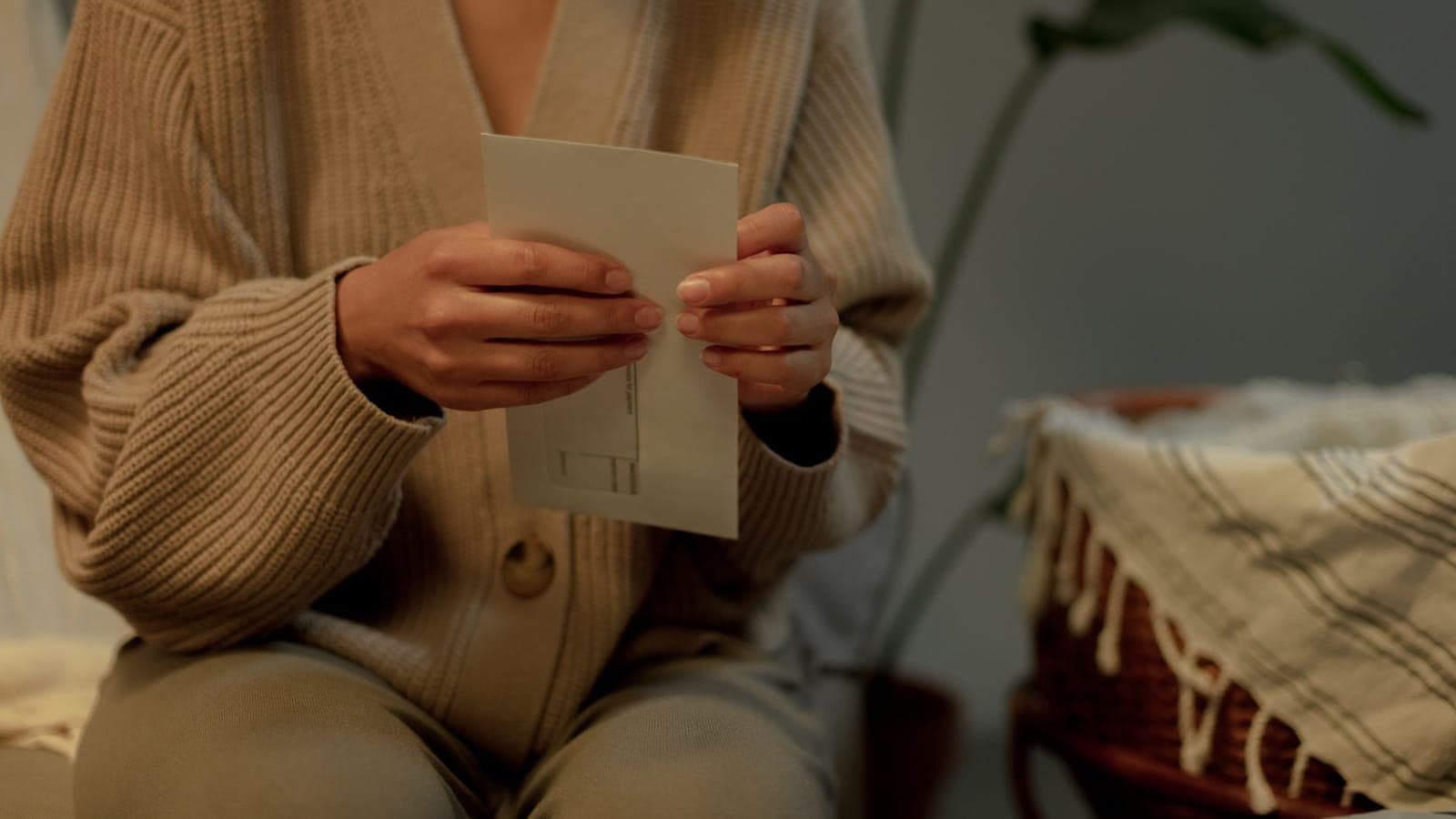 Two hands hold a white window envelope in front of a sand-coloured cardigan; on the right, a wicker basket covered with a striped throw
