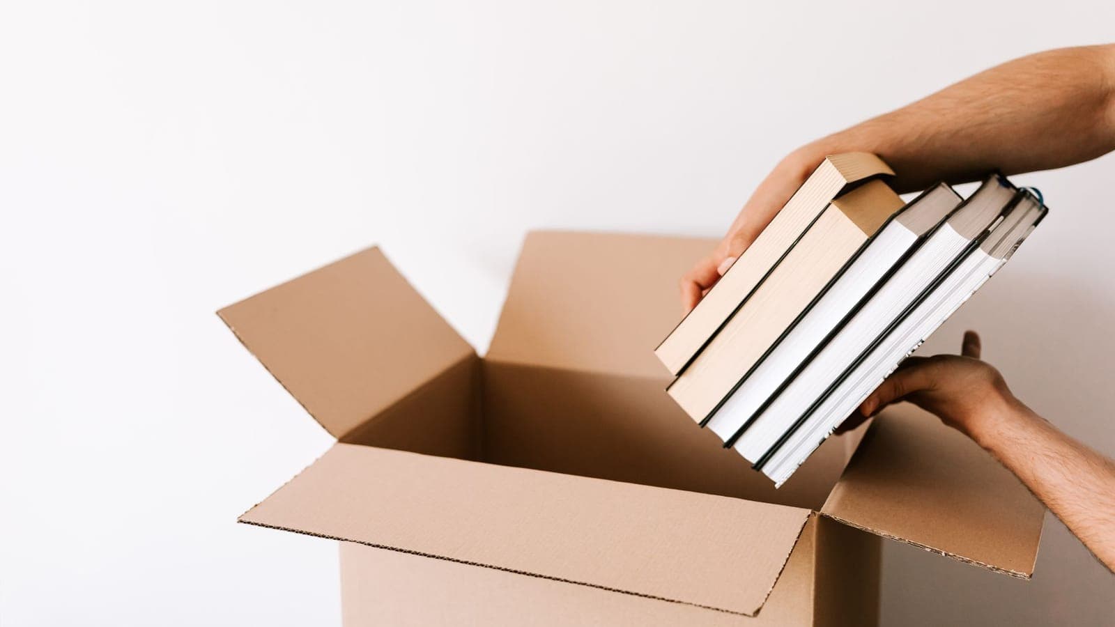 Crop unrecognizable male packing staked books into carton box while standing against white wall during relocation