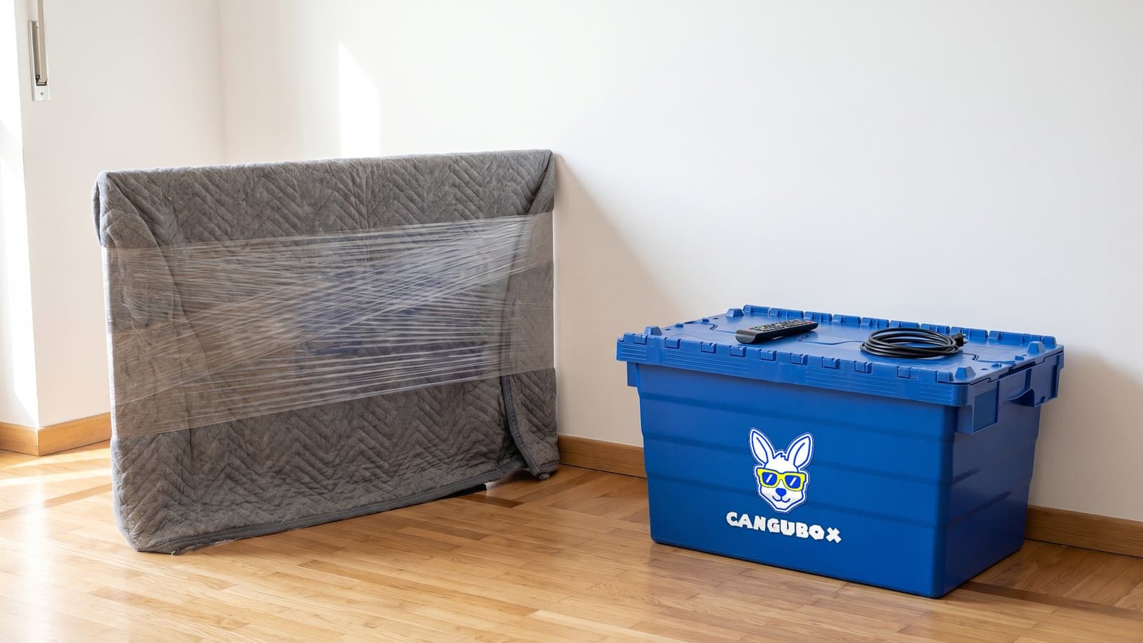 A television wrapped in a grey quilted blanket held by stretch wrap, standing against a white wall next to a closed blue Cangubox with a remote control and a coiled cable on its lid