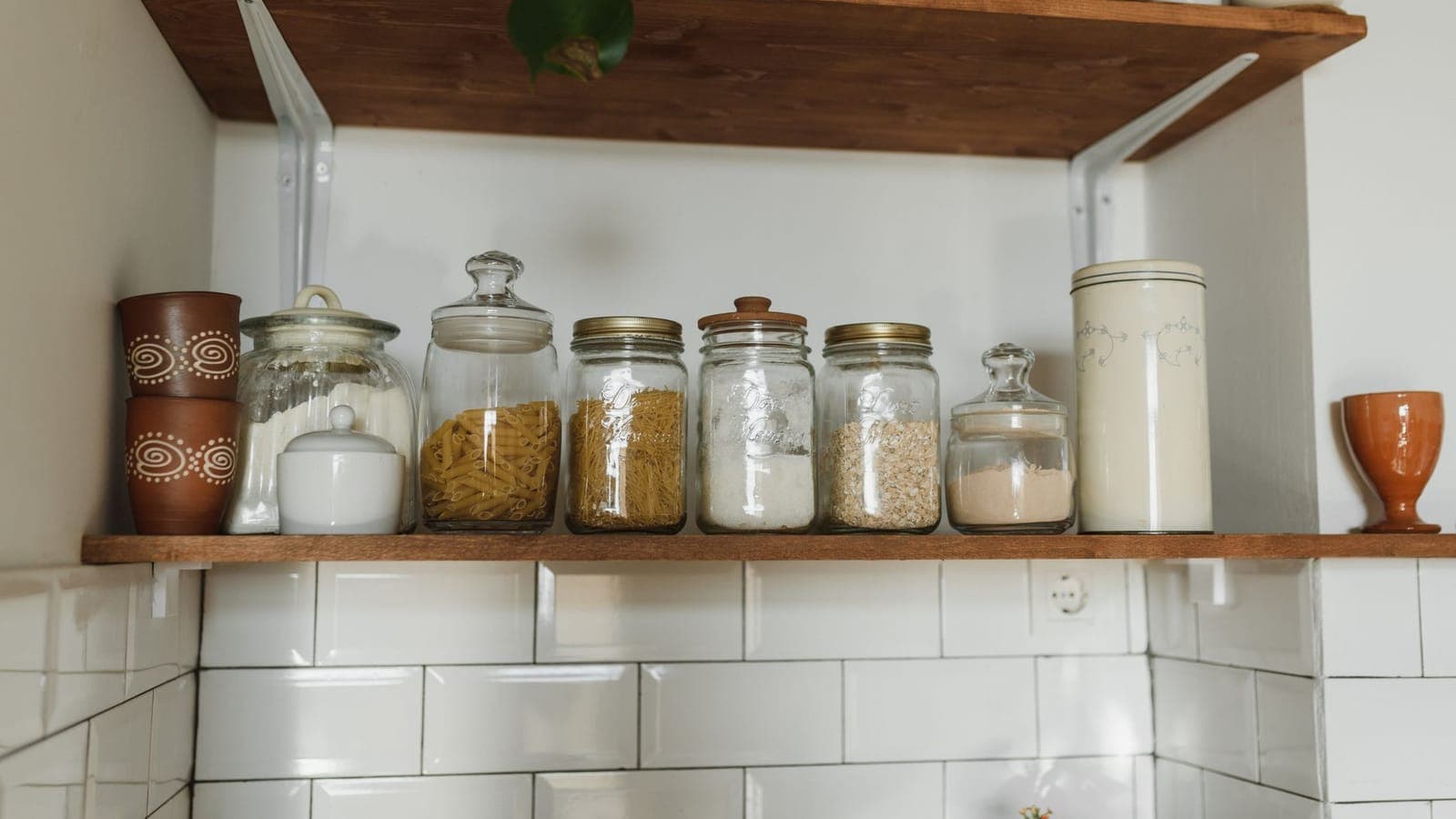 Una mensola di legno sopra le piastrelle bianche di una cucina, con una fila di barattoli di vetro pieni di pasta, fiocchi d'avena, zucchero e farina, fra due tazze di terracotta e un barattolo di latta color crema