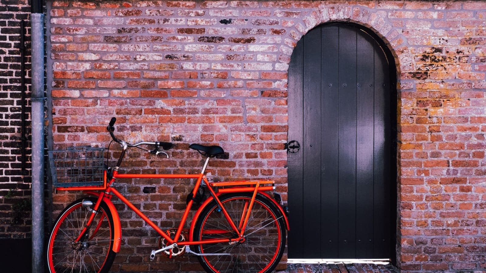 Una bicicletta rossa con il cestino davanti e il portapacchi dietro, appoggiata a un muro di mattoni accanto a una porta nera ad arco
