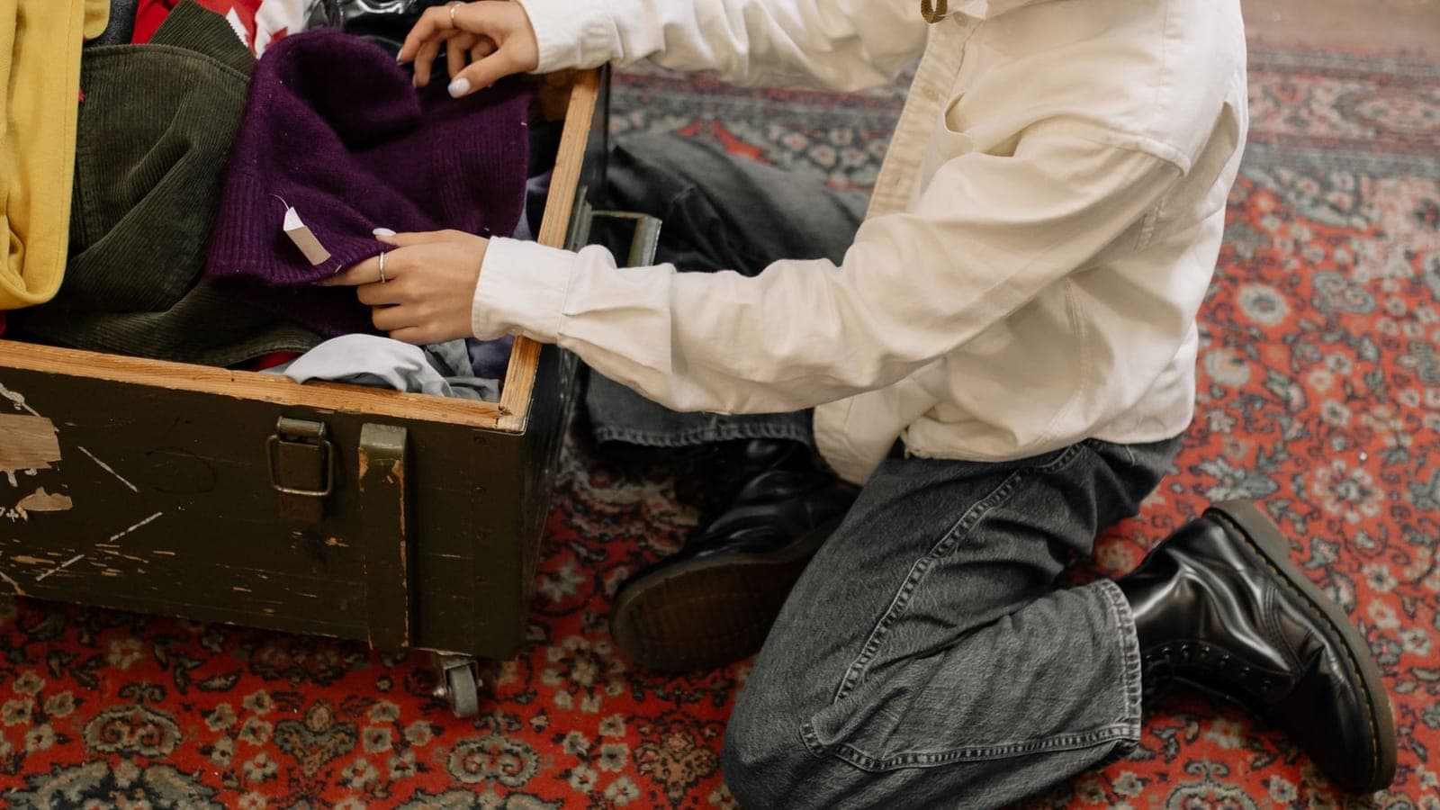 A person kneeling on a red patterned carpet rummages inside a wooden trunk full of jumpers and clothes piled up
