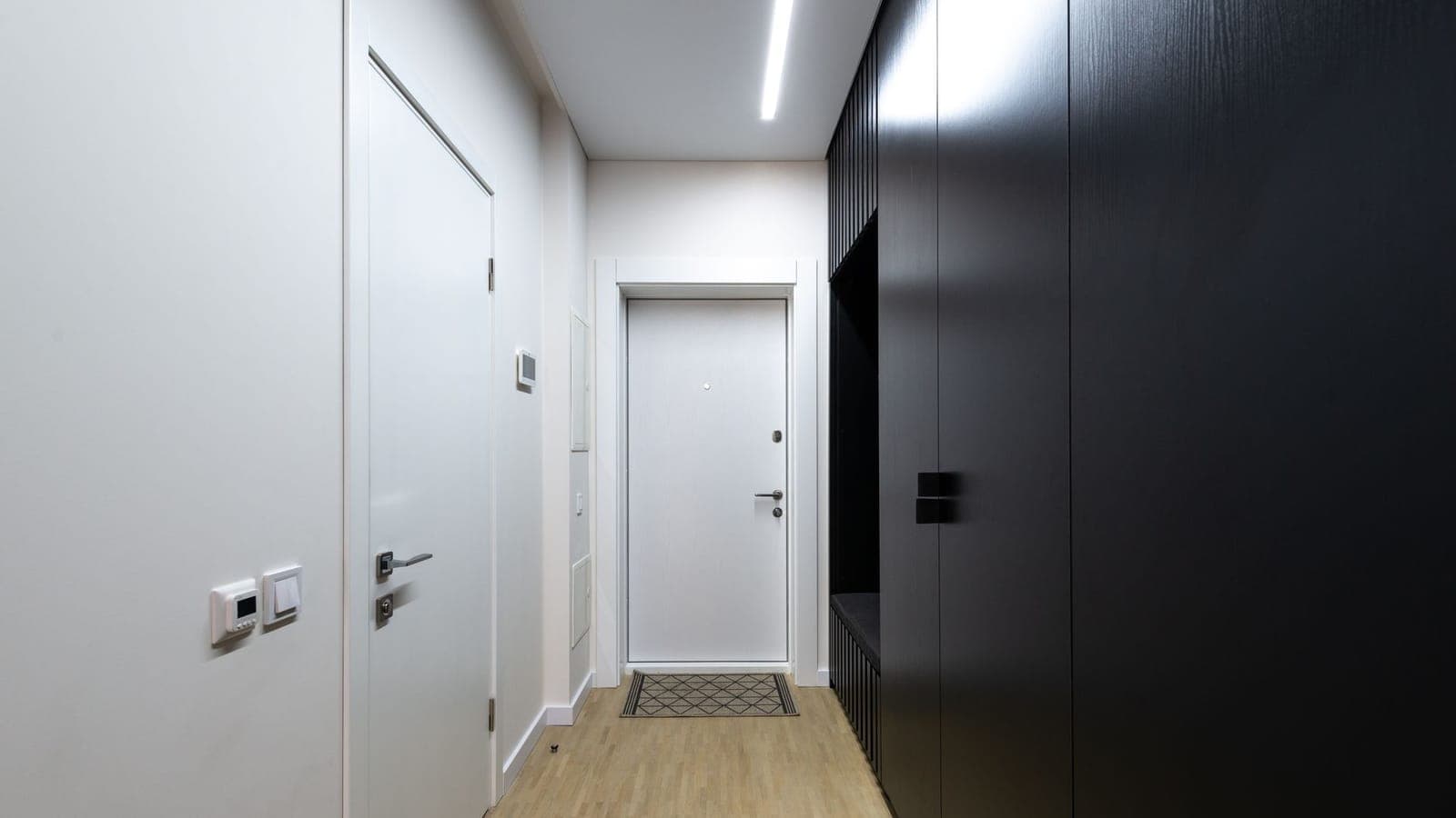 Un corridoio d'ingresso vuoto con il pavimento in legno chiaro: in fondo la porta di casa bianca e chiusa con uno zerbino davanti, a destra una parete di armadi neri, a sinistra una porta bianca