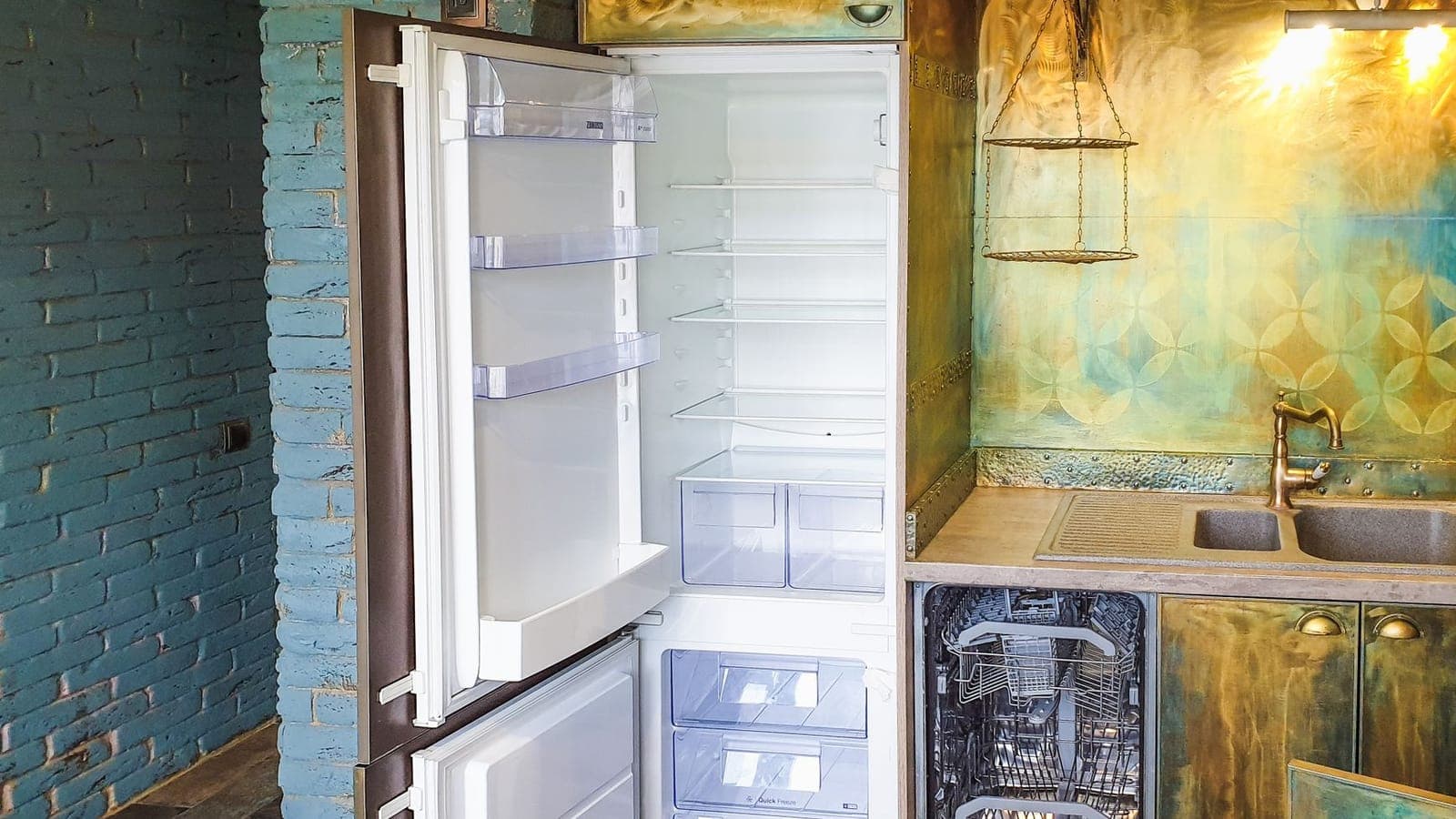 Un frigorifero da incasso aperto e completamente vuoto, coi ripiani di vetro a vista, accanto a una lavastoviglie aperta in una cucina dalle ante di ottone e dal muro di mattoni azzurri