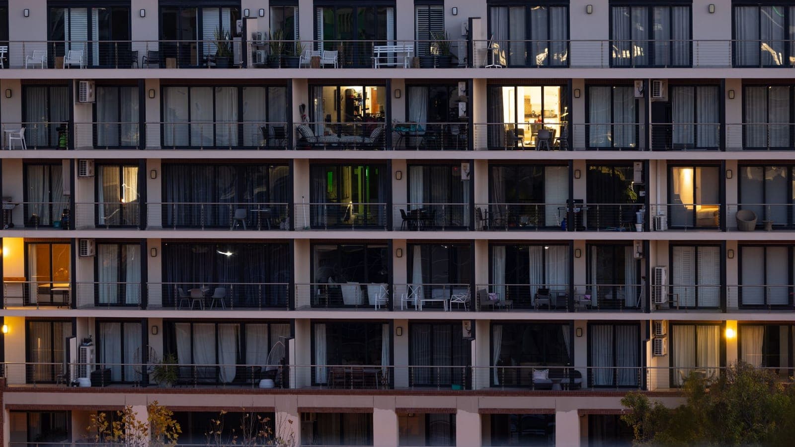 La facciata di un condominio all'imbrunire, con file di balconi e tende tirate: solo poche finestre sono accese, fra cui una cucina con la luce calda e un soggiorno con un divano a righe sul balcone