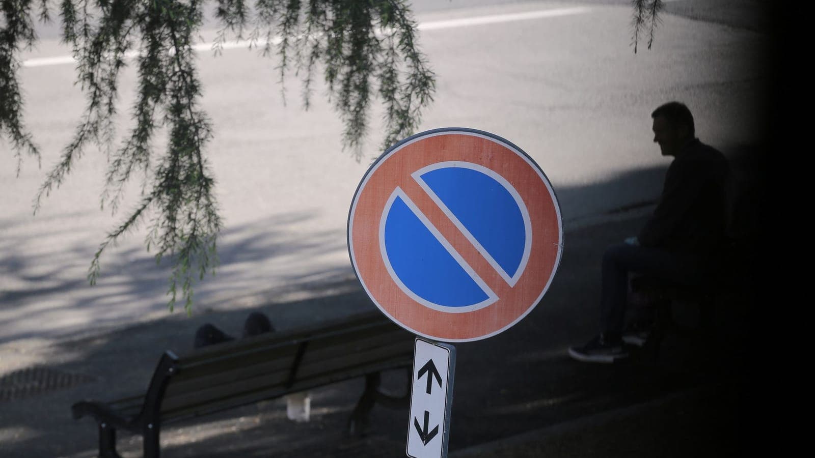 A no parking sign with its arrow plate, in front of an empty road, with a bench and a man sitting in silhouette