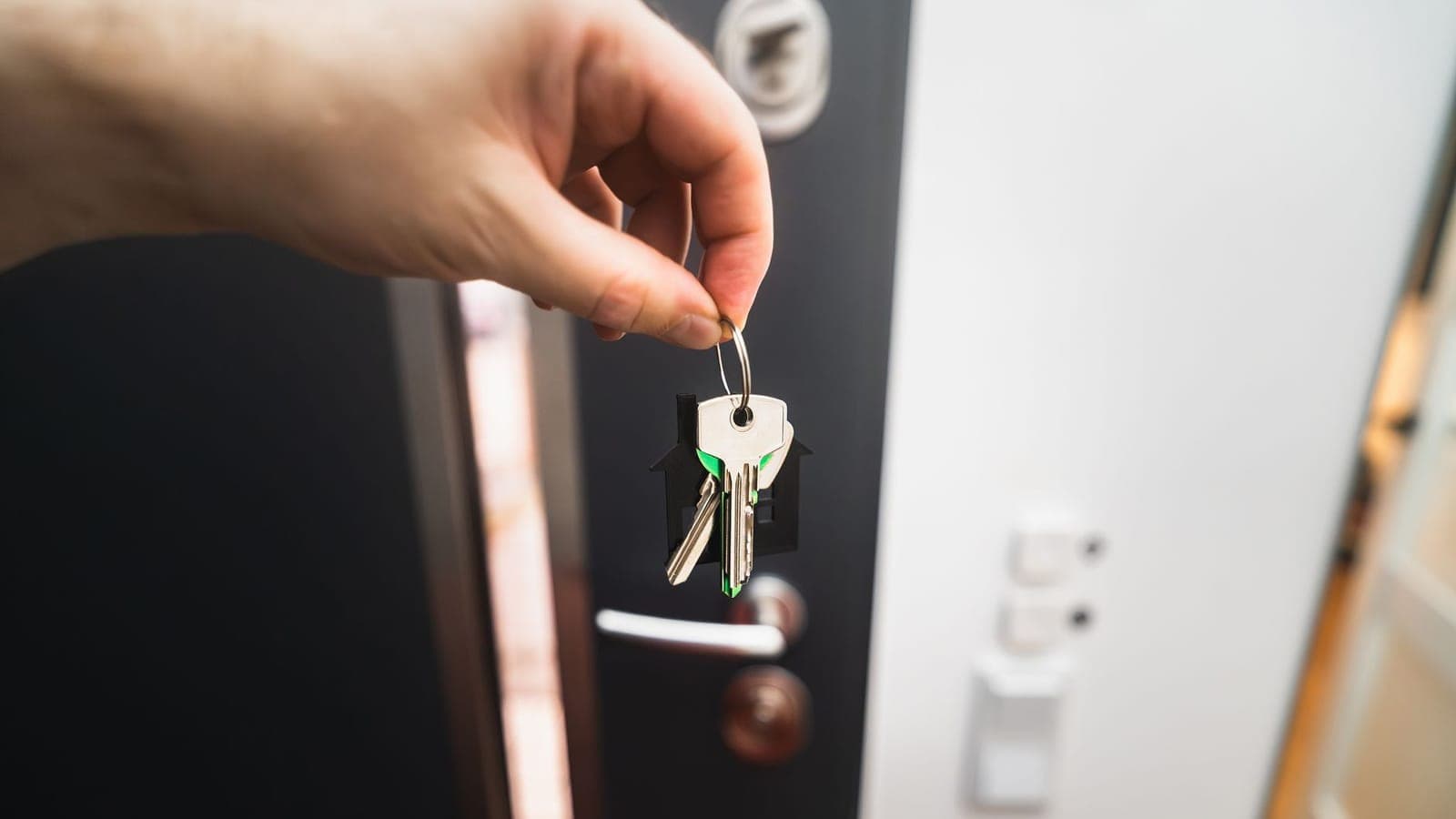 A hand holding two keys on a green and black house shaped keyring, in front of an open dark front door, with a white wall and light switches blurred behind