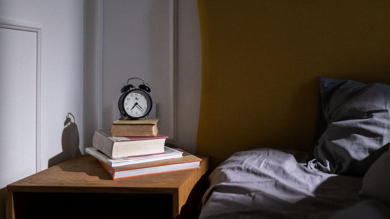 Una sveglia a campanelle nera appoggiata su una pila di libri, sul comodino di legno di una camera da letto, accanto a un letto disfatto e a una parete color senape
