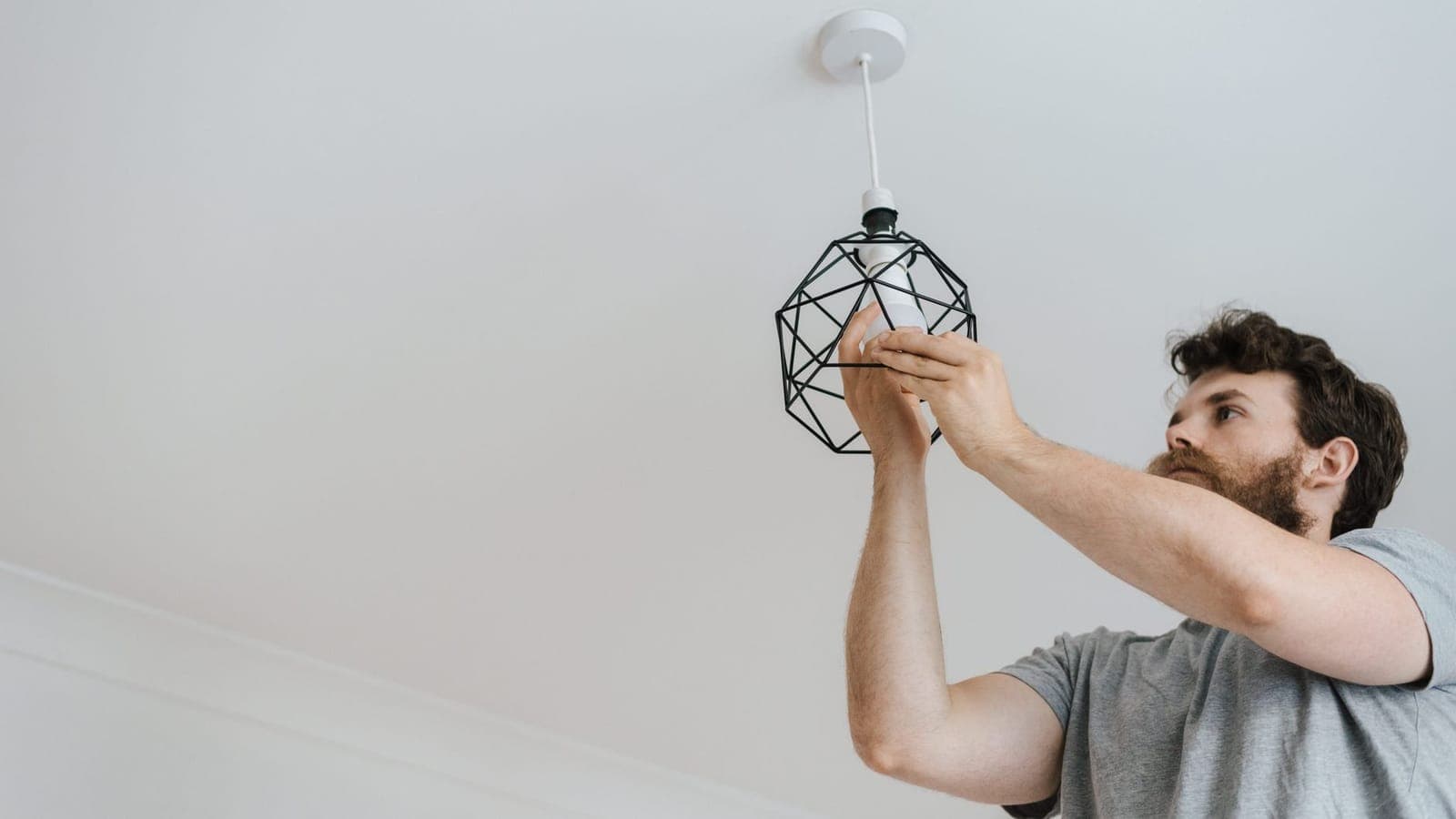 Un uomo con la barba e la maglietta grigia, visto dal basso, tiene con due mani la lampadina dentro una lampada a gabbia nera appesa a un soffitto bianco