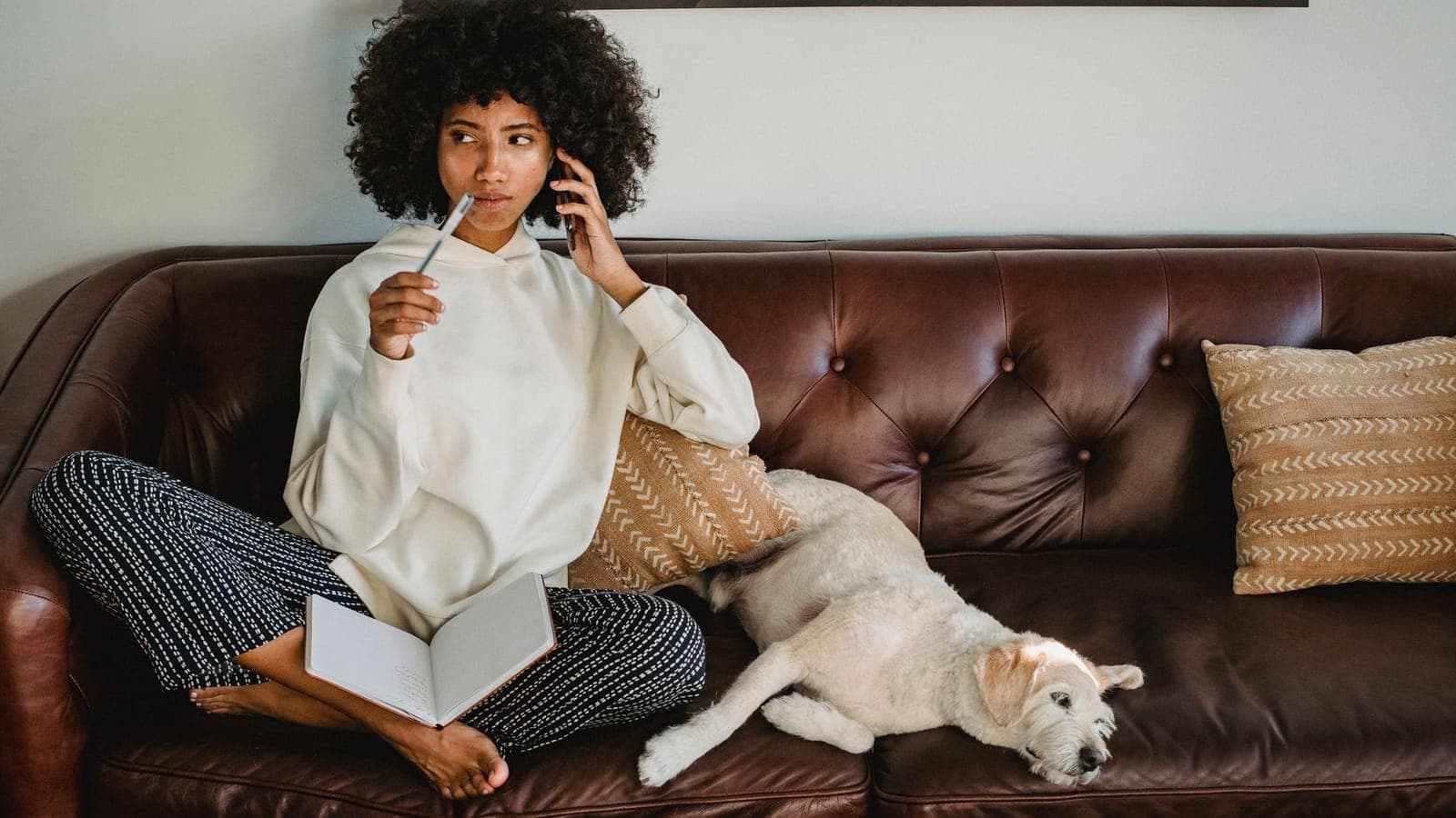 A young woman in a white sweatshirt, sitting cross-legged on a brown leather sofa, talks on the phone with a pen in her hand and an open notebook on her knees; next to her a light-coated dog is asleep