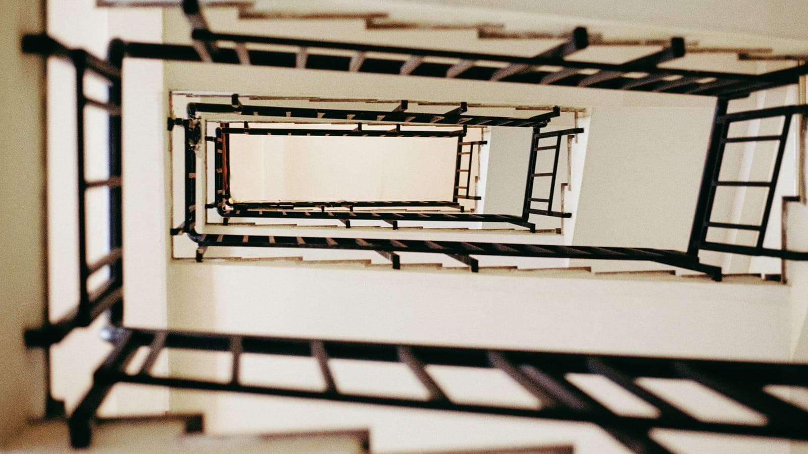 The stairwell of a block of flats seen from below, flights rising one above the other around a rectangular well, black railings and cream walls