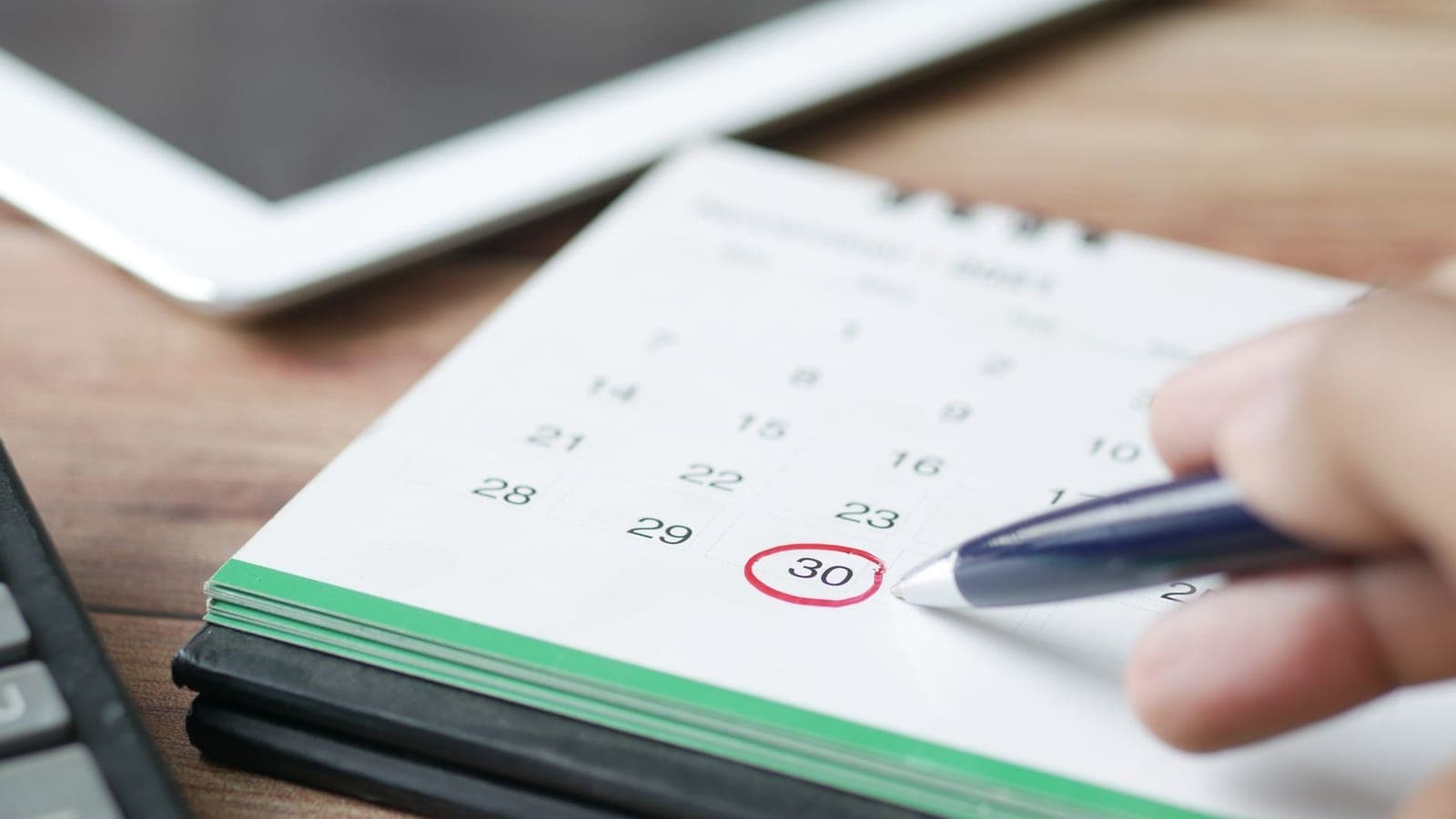 A spiral bound desk calendar on a wooden desk, with the number thirty circled in red and a hand resting a dark blue pen on it; behind, out of focus, a white tablet and the corner of a keyboard